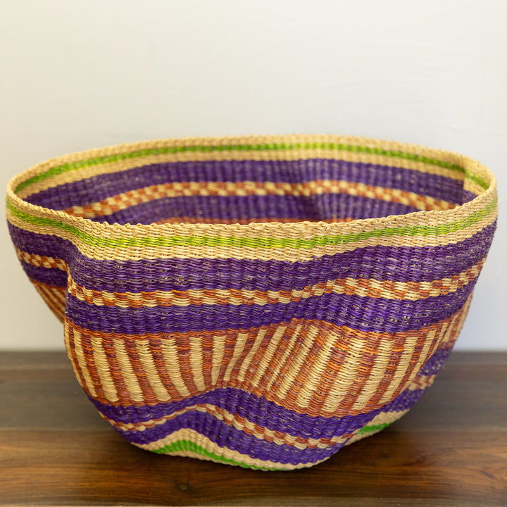 Woven basket with purple and green stripes on a wooden surface, handwoven fair trade wavy Bolga basket with leather handle.


