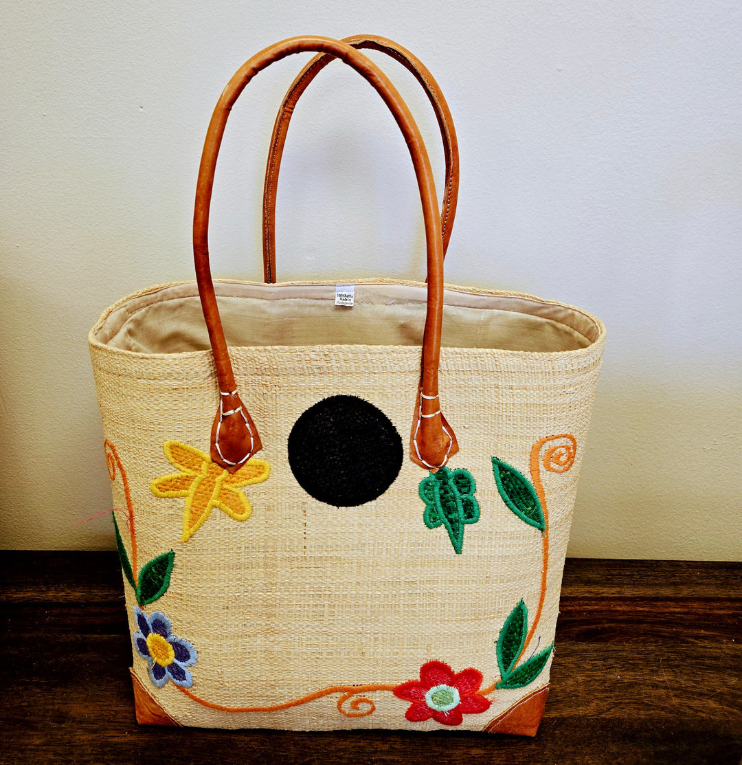 Handwoven raffia handbag with colorful floral design and leather handles.