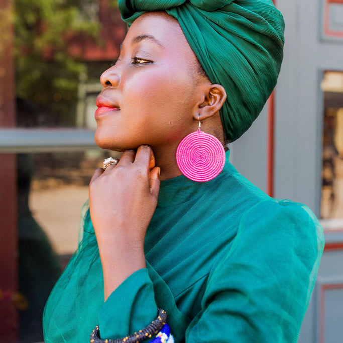 Woman wearing a green headscarf and raffia pink earrings, standing in front of a building, enjoying the summer. Earrings perfect for a summer day.