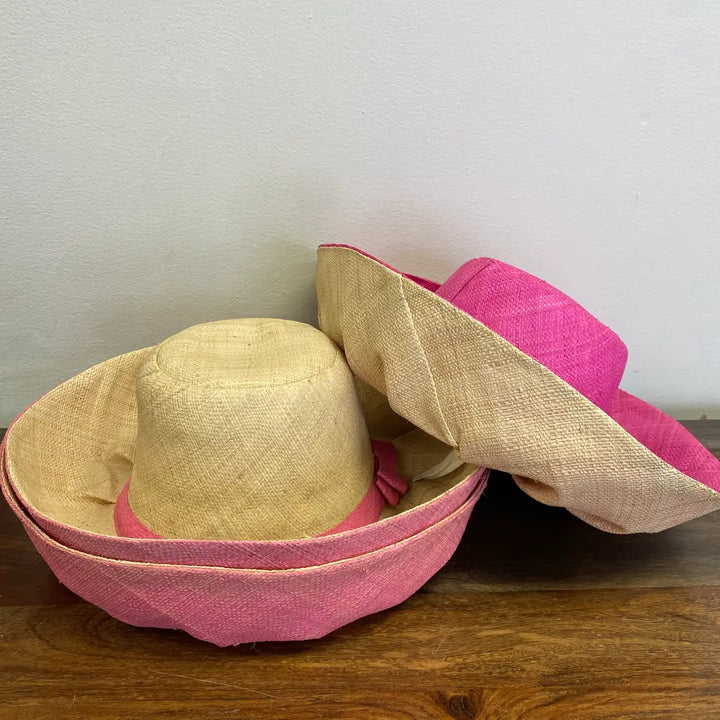 Two straw hats, one pink and one beige, on a wooden surface with a white wall background.