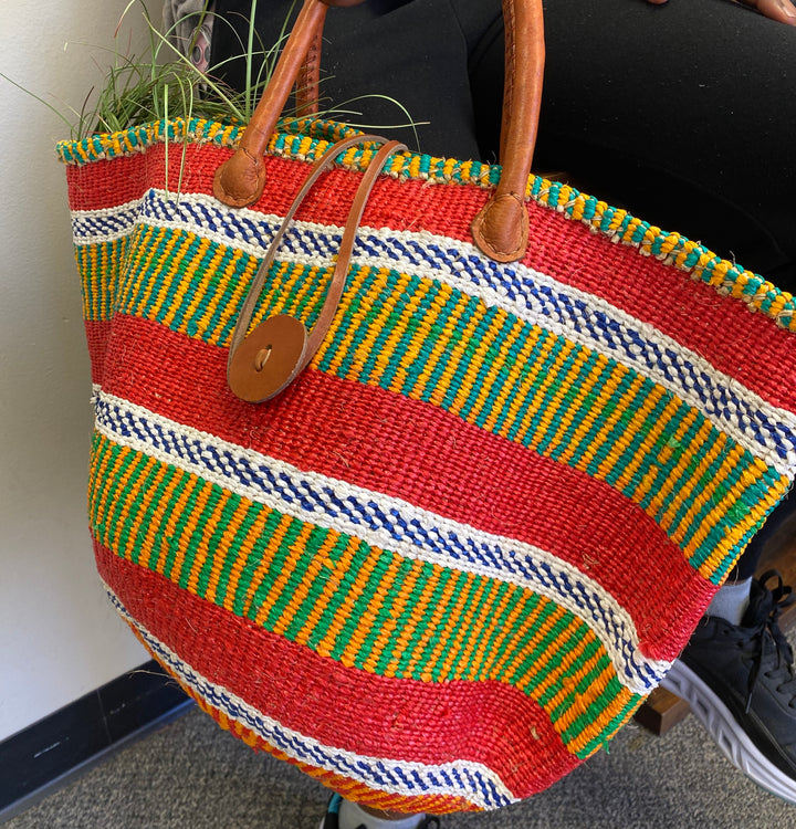 Colorful striped woven bag with brown leather handles on a neutral background.