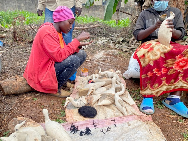 Artisans in Kenya sitting outdoors on the ground carving wooden bird figurines and other handcrafted items laid out on a blanket in their workshop.