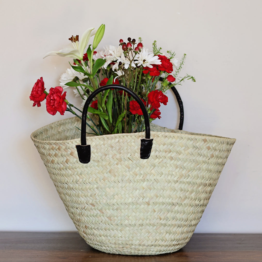 Woven straw tote bag with black leather handles holding a bouquet of flowers, perfect for an everyday market basket
