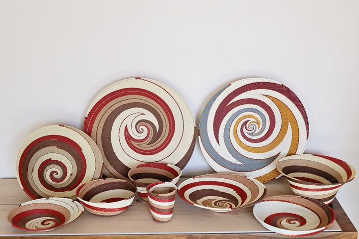 A collection of Zulu hand-woven spiral-patterned ceramic tableware in our Desert design, including plates, bowls, and a small cup, arranged on a wooden surface against a plain white wall.