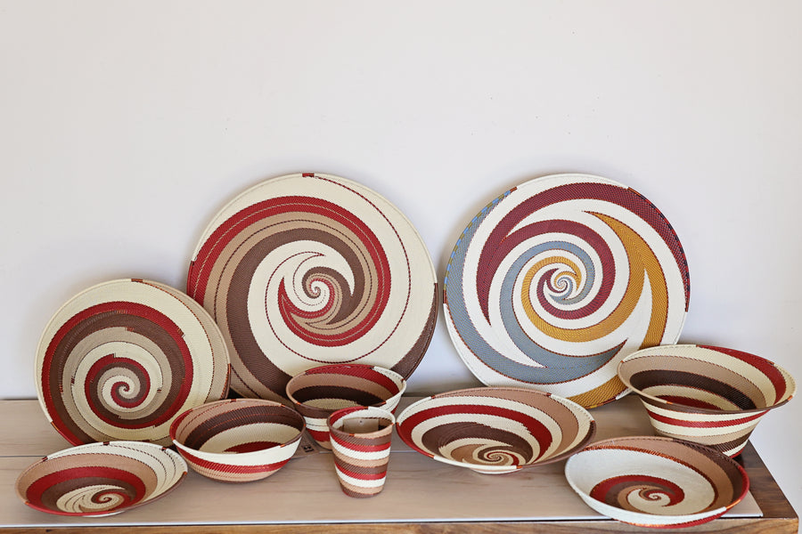 A collection of Zulu hand-woven spiral-patterned ceramic tableware in our Desert design, including plates, bowls, and a small cup, arranged on a wooden surface against a plain white wall.