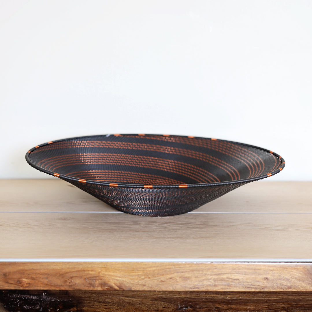 A wide, shallow woven bowl with a black exterior and copper-brown striped interior, displayed on a light wood surface against a plain white wall.