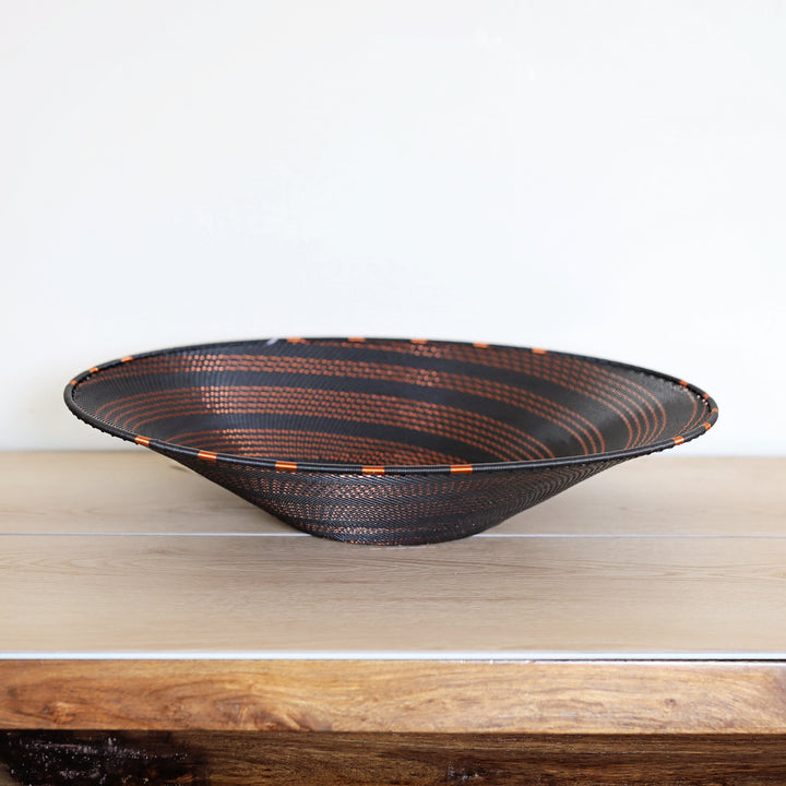 A wide, shallow woven bowl with a black exterior and copper-brown striped interior, displayed on a light wood surface against a plain white wall.