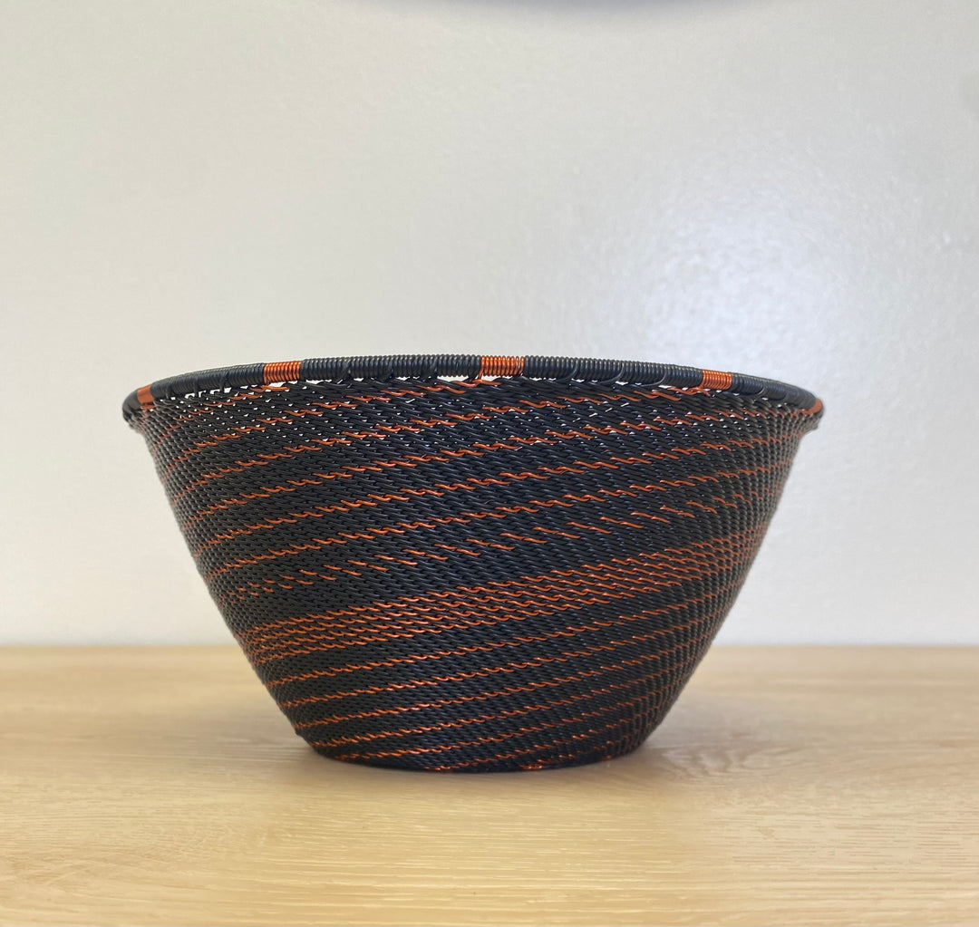 A hand-woven deep basket bowl with a black and orange striped pattern, sitting on a light wood surface against a plain off-white wall.