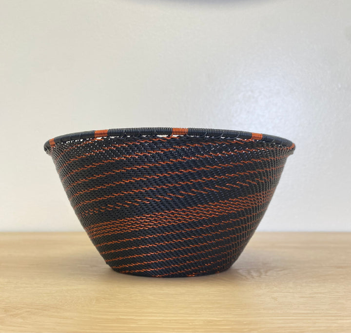 A hand-woven deep basket bowl with a black and orange striped pattern, sitting on a light wood surface against a plain off-white wall.