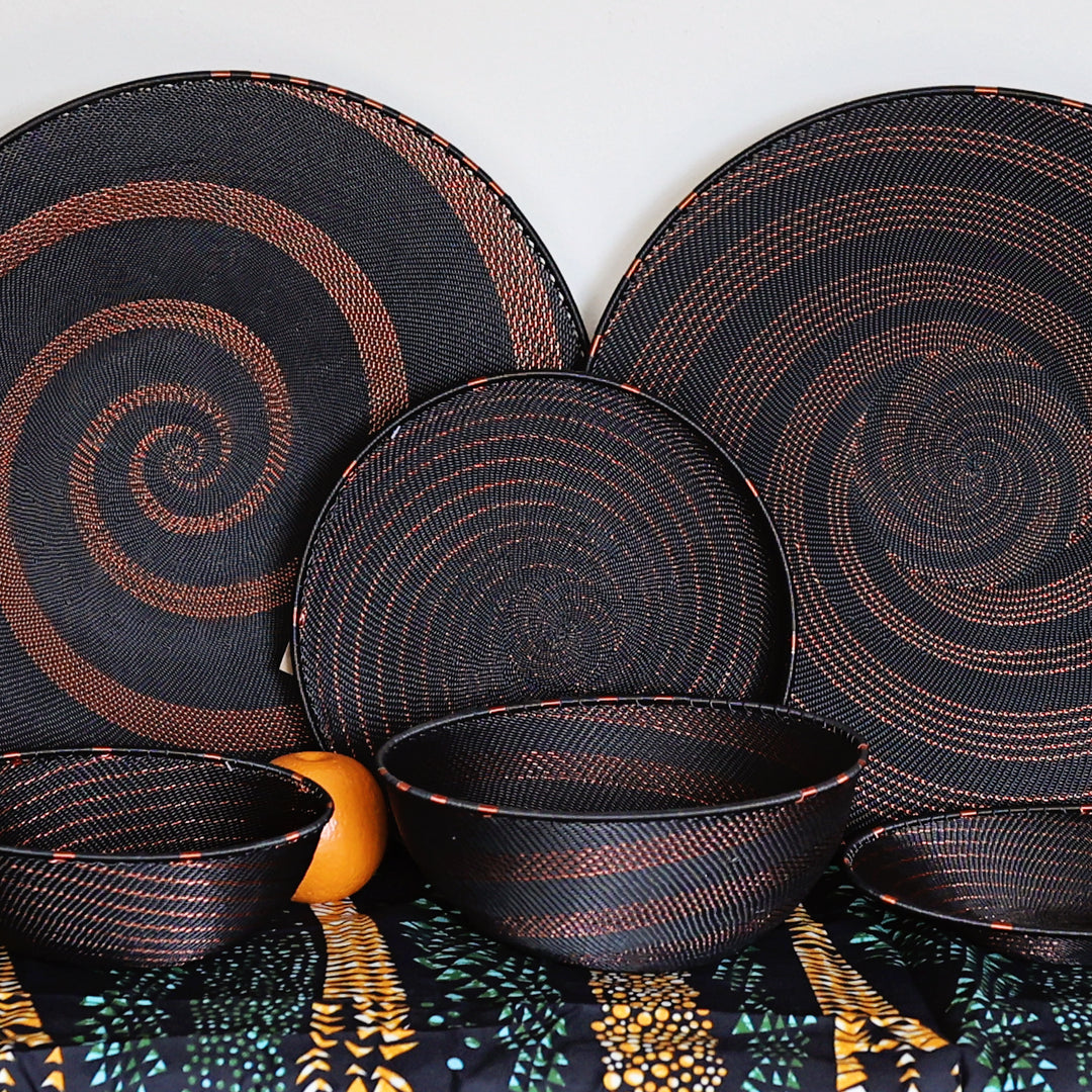 A coordinated set of woven black tableware with copper-brown spiral patterns, including two large round platters, two round plates, two bowls, and a patterned table runner, arranged against a plain light background with a small orange fruit and green decorative accents.