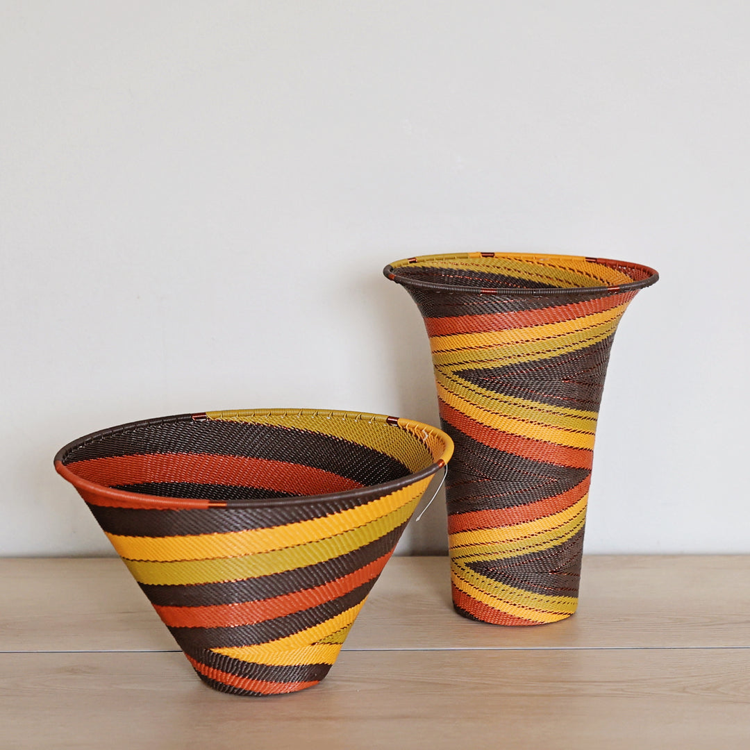 Two hand-woven basket vessels with diagonal striped patterns in yellow, orange, red, and dark brown, displayed on a light wood surface against a plain white wall.