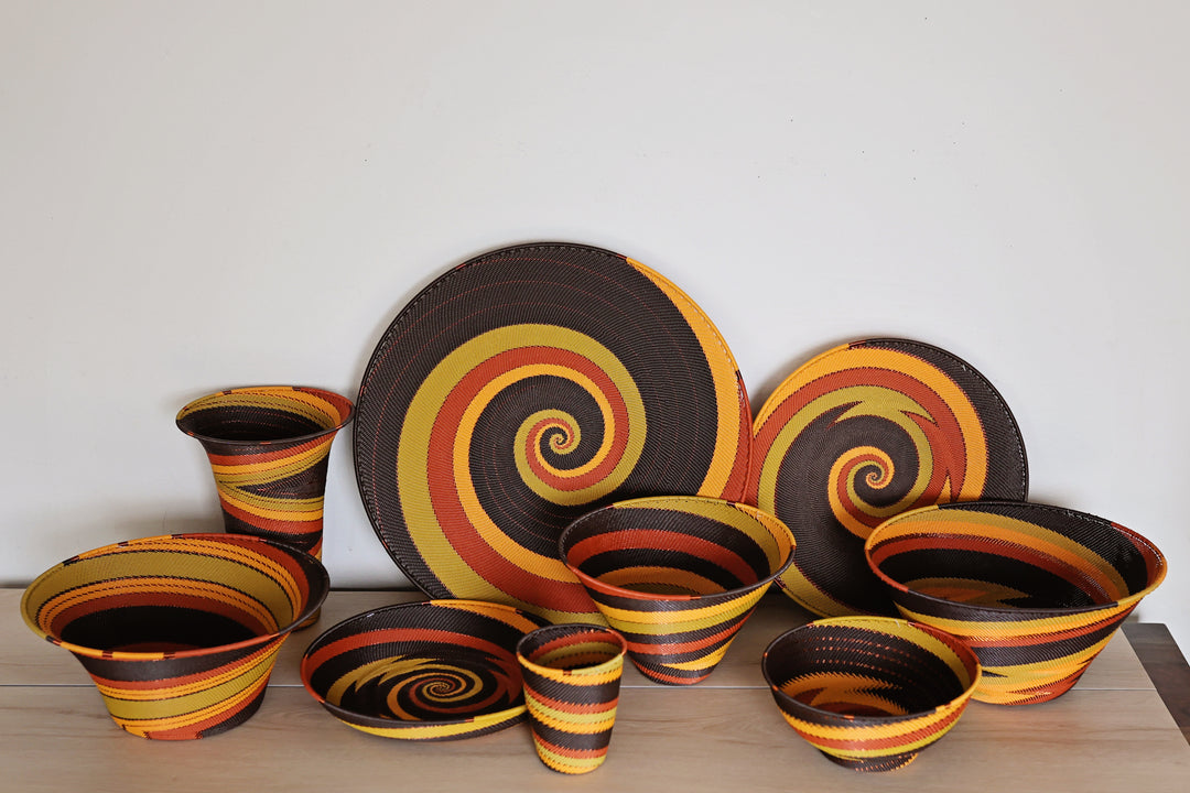 A coordinated set of Dark Earth colored Zulu handwoven baskets in a bold spiral pattern of dark brown, yellow, orange, and red, displayed on a light wood surface against a plain white wall.