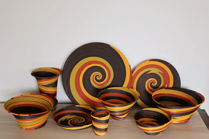 A coordinated set of Dark Earth colored Zulu handwoven baskets in a bold spiral pattern of dark brown, yellow, orange, and red, displayed on a light wood surface against a plain white wall.
