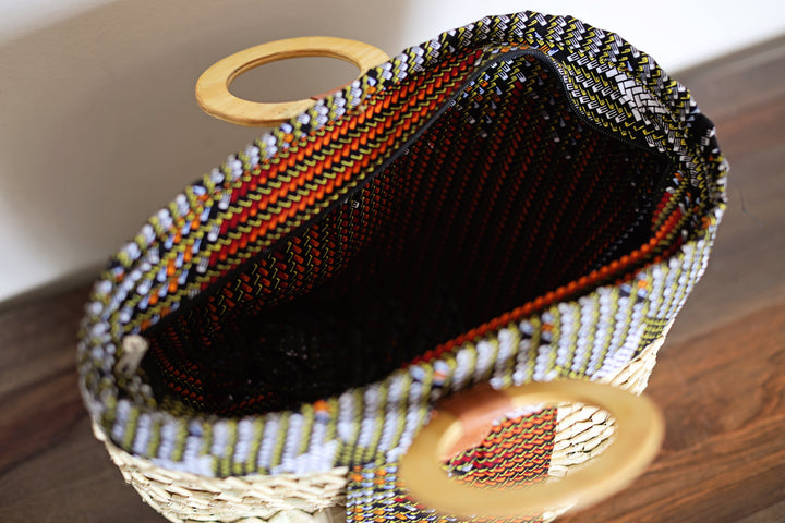 A close-up of a woven handbag with a black interior, featuring a patterned exterior of white, green, and orange woven threads, and two round light wood handles on a wooden table.