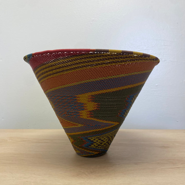 A hand-woven large deep funnel zulu bowl with a vibrant geometric pattern in red, yellow, orange, brown, and blue, displayed on a light wood surface against a plain white wall.