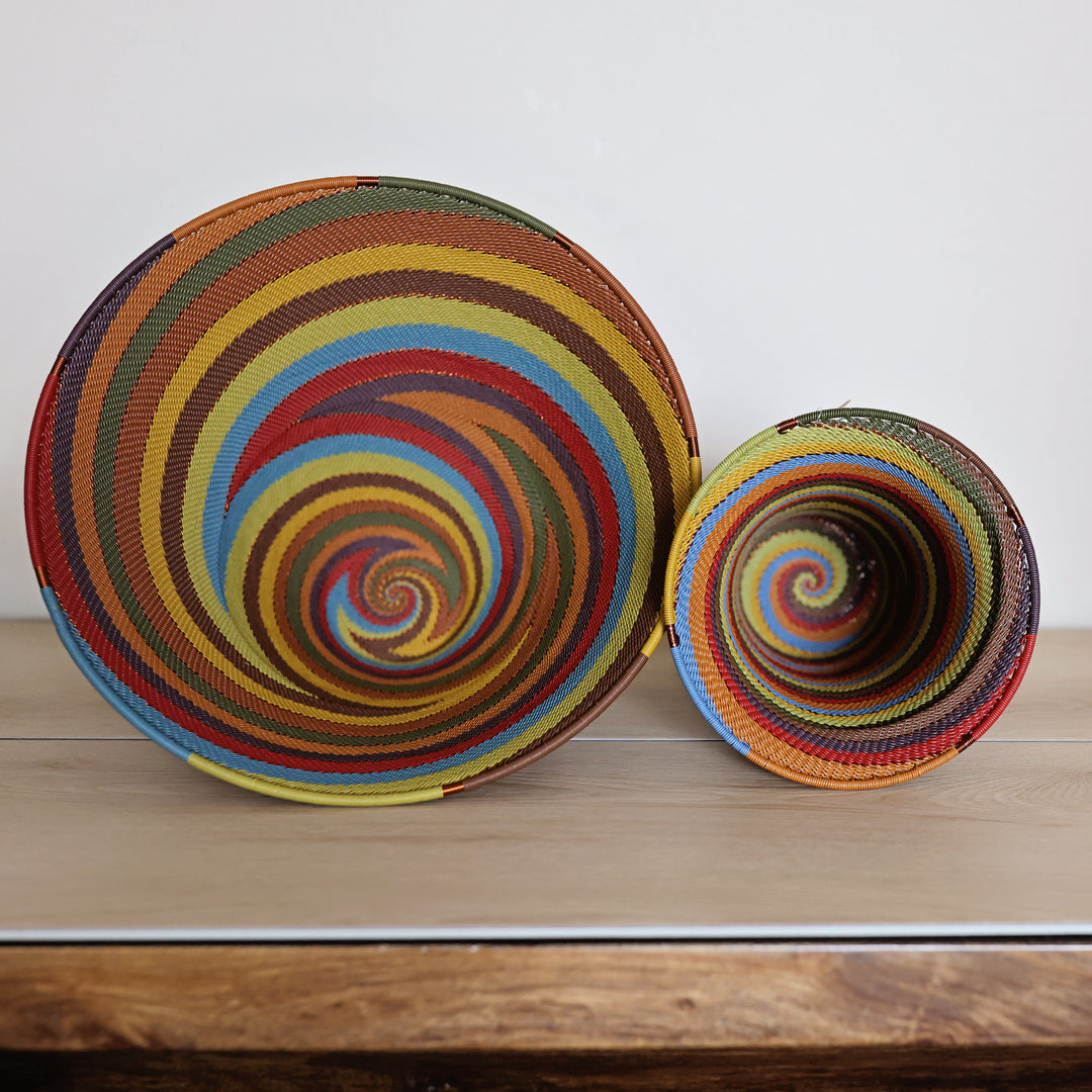 Two hand-woven Zulu spiral baskets in vibrant multicolor stripes, one standing upright and the other tilted, displayed on a light wooden surface against a plain white wall.