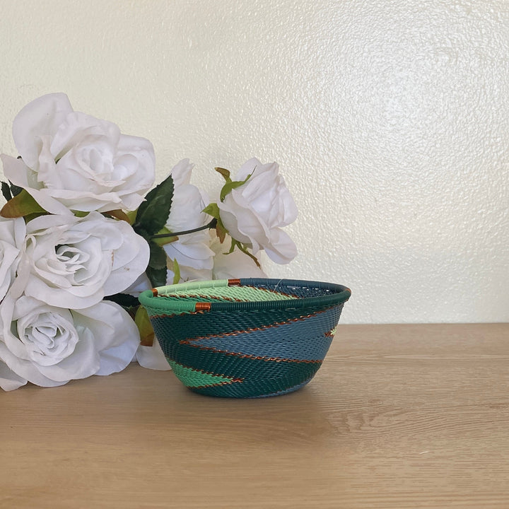 A small handwoven emerald zulu basket bowl with a green and blue pattern sits on a light wood table next to white faux roses, with a cream wall and a bamboo sunburst mirror in the background.