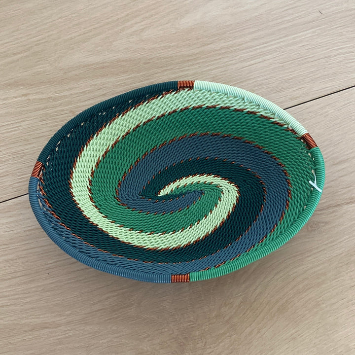A hand-woven oval emerald basket with a spiral pattern of green, teal, and light mint, resting on a light wood floor.