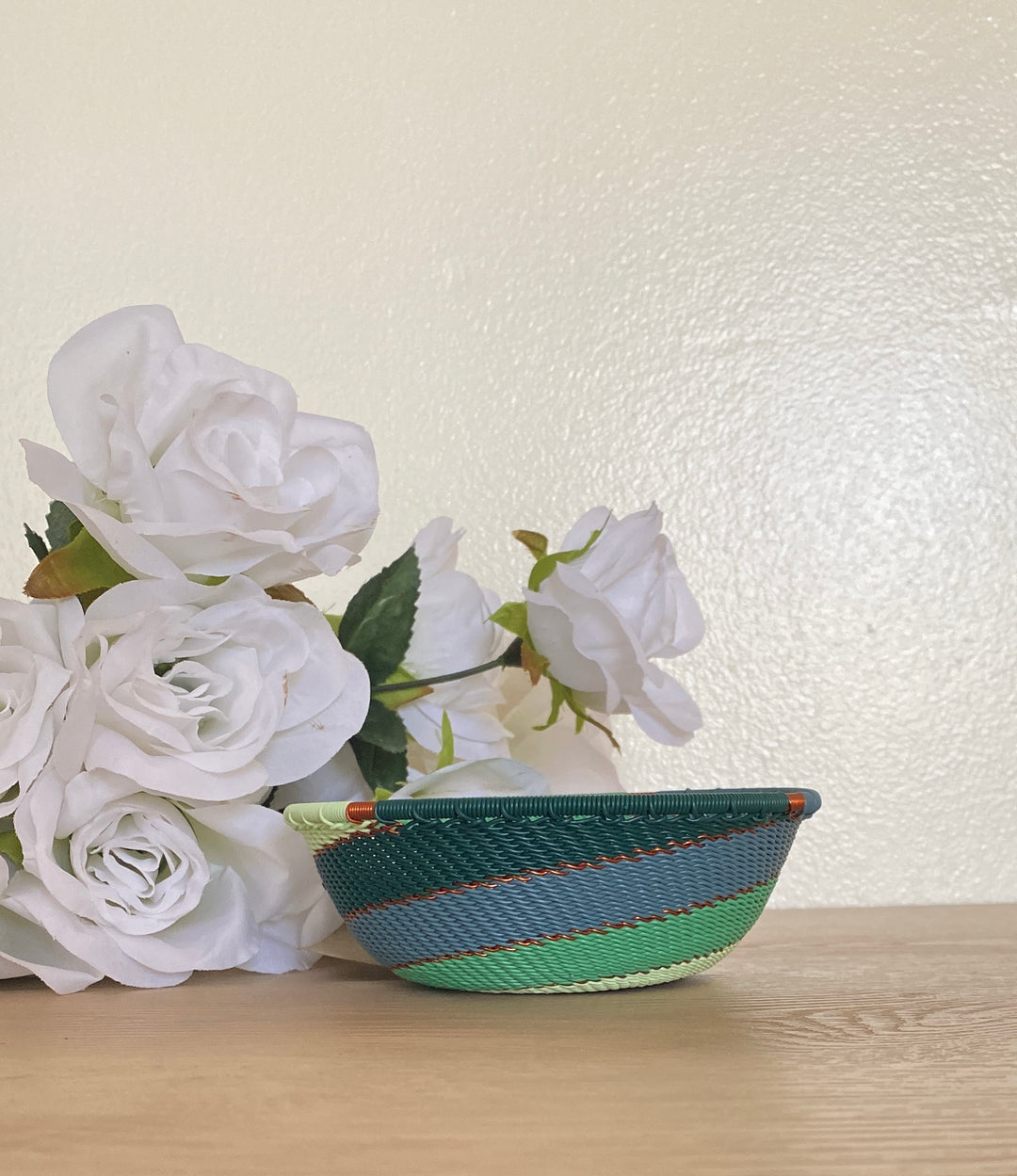 A small woven emerald zulu basket bowl with green, blue, and orange stripes sits on a light wood surface next to white faux roses against a textured cream wall.
