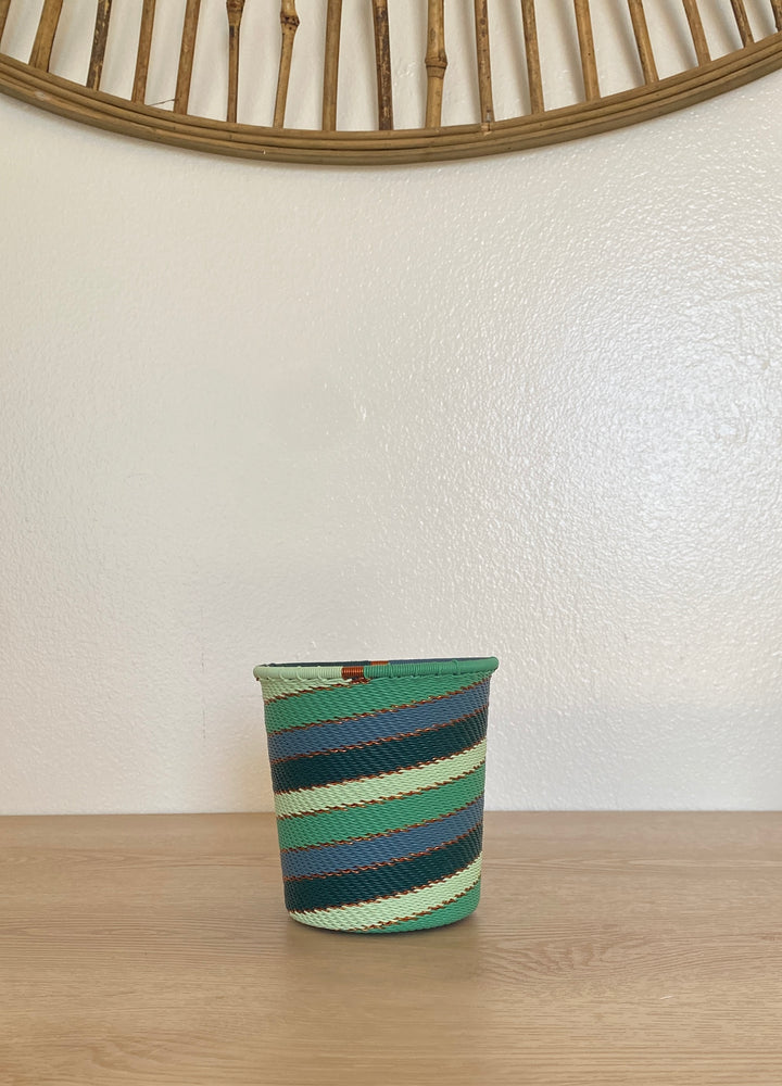 A small woven zulu emerald basket with horizontal stripes in green, teal, navy, and cream, sitting on a light wood tabletop against a white wall.