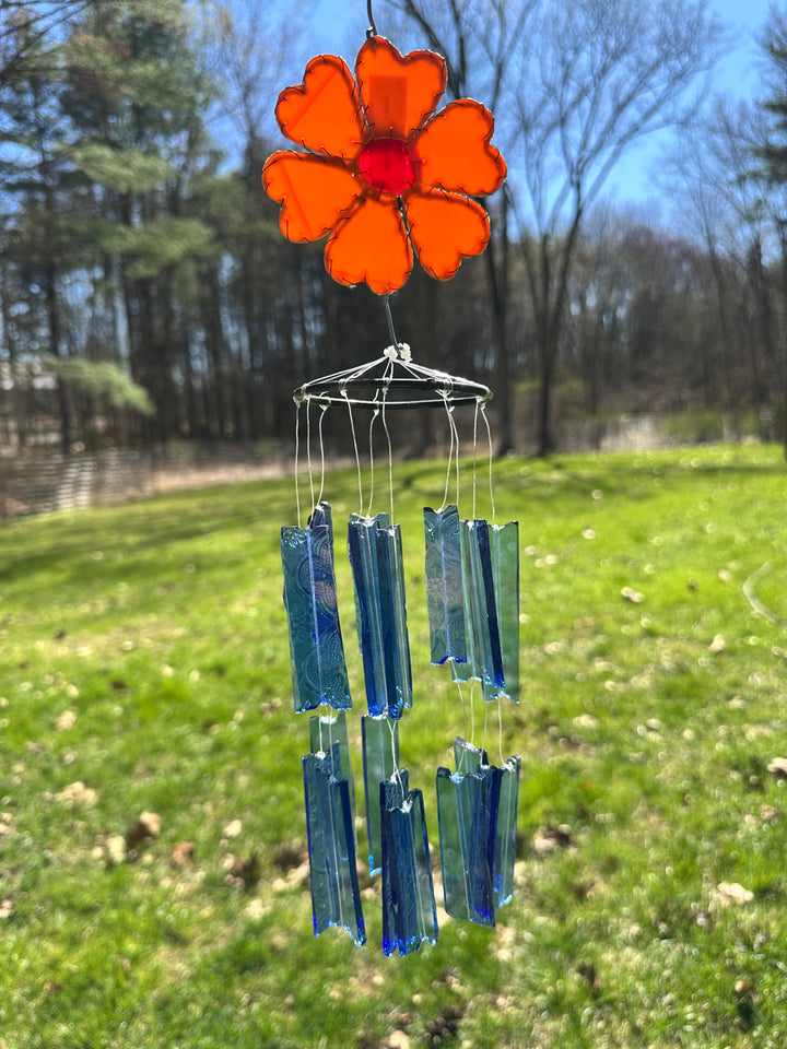 Decorative glass wind chime with an orange flower and blue glass rectangular pieces in a garden setting.