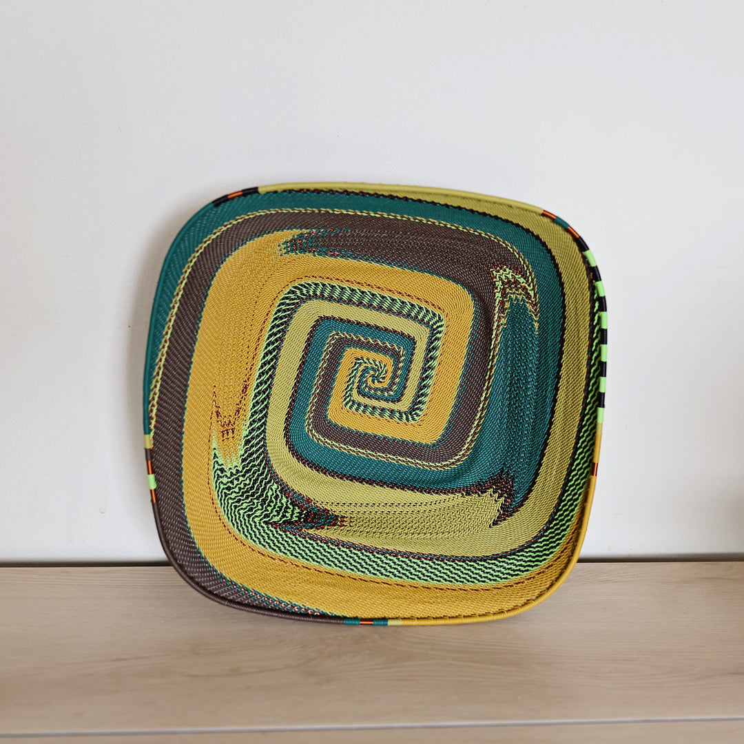 A square woven basket with rounded corners, featuring a spiral pattern in yellow, teal, brown, and green, resting on a light wood surface against a white wall.
