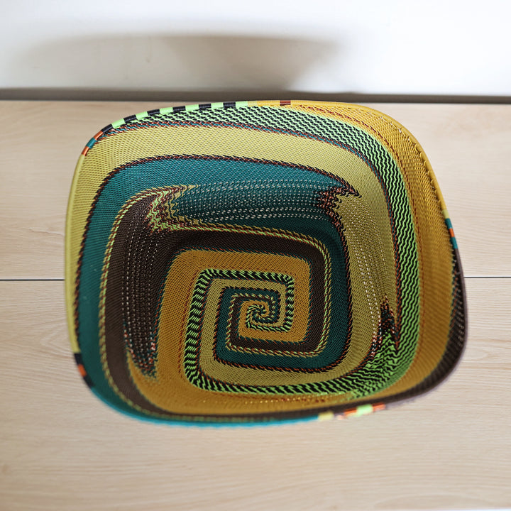 A hand-woven oval basket with a vibrant spiral pattern in yellow, green, teal, brown, and black, photographed from directly above on a light wood floor.