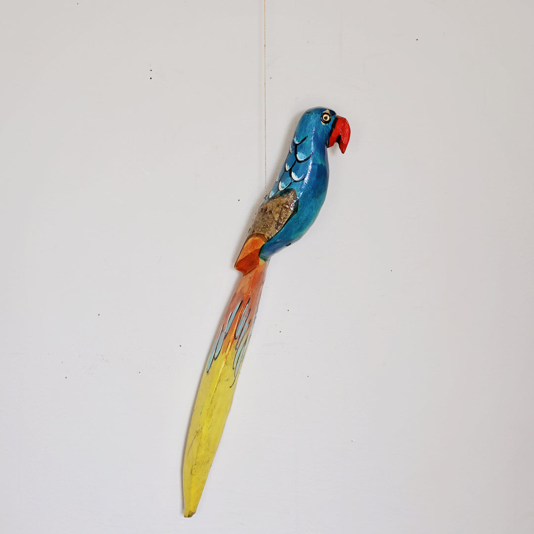 A colorful hand-painted wooden wall hanging shaped like a parrot, featuring a blue head and back, a red beak, orange wings, and a long yellow tail, hanging from a string.