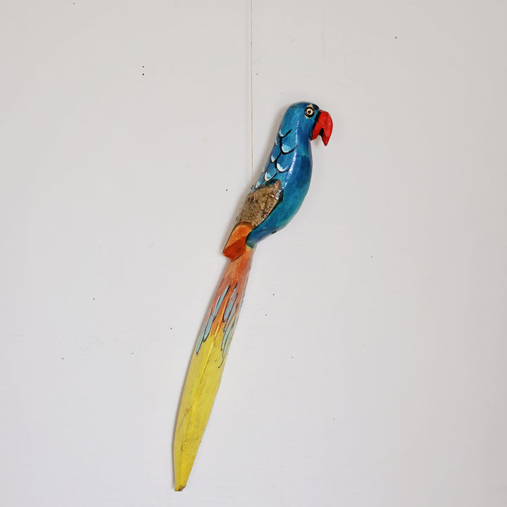 A colorful hand-painted wooden wall hanging shaped like a parrot, featuring a blue head and back, a red beak, orange wings, and a long yellow tail, hanging from a string.