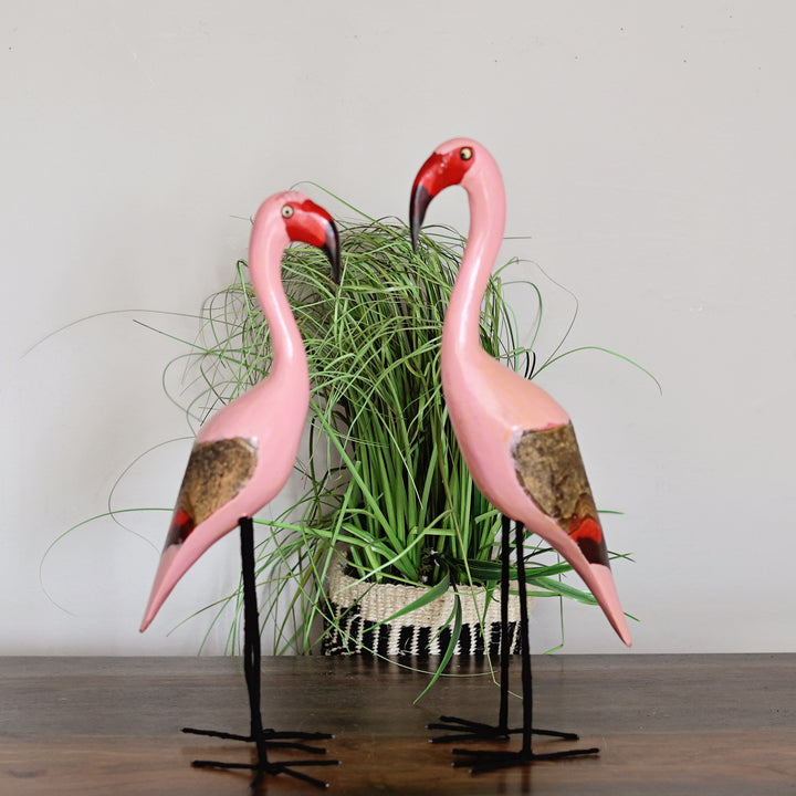 Two handcrafted, pink flamingo figurines with red beaks and brown patterned wings, displayed on black metal stands in front of a potted green plant against a plain light gray wall.