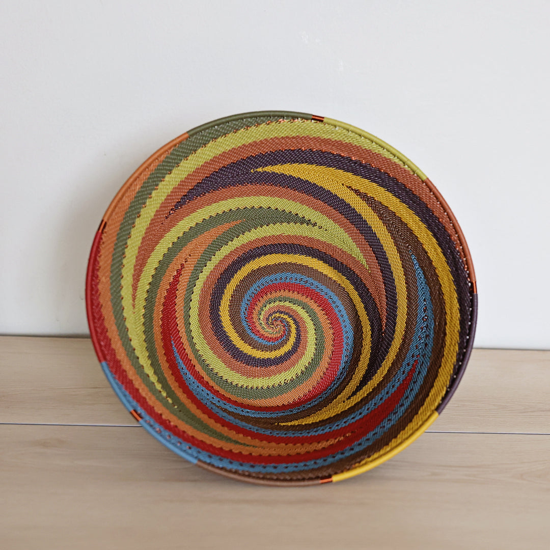 A round woven handwoven zulu basket in Earthy Rainbow color with a vibrant rainbow spiral pattern, photographed standing on a light wood surface against a plain white wall.