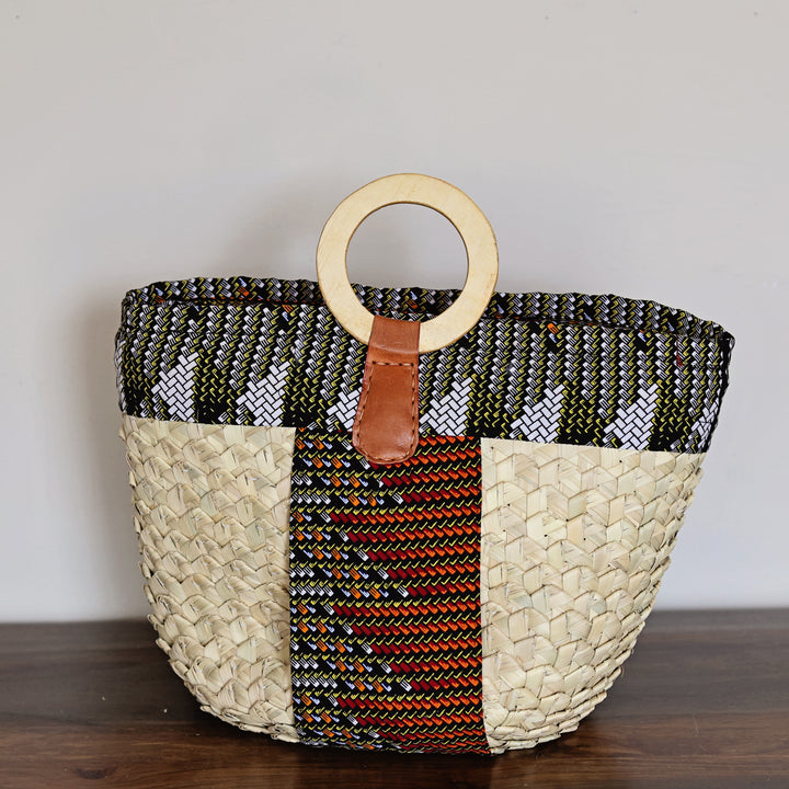 A woven tote bag with a natural straw body, a black and white patterned upper band, a central red and black patterned stripe, and a light wood ring handle, resting on a dark wooden surface against a plain light gray wall.