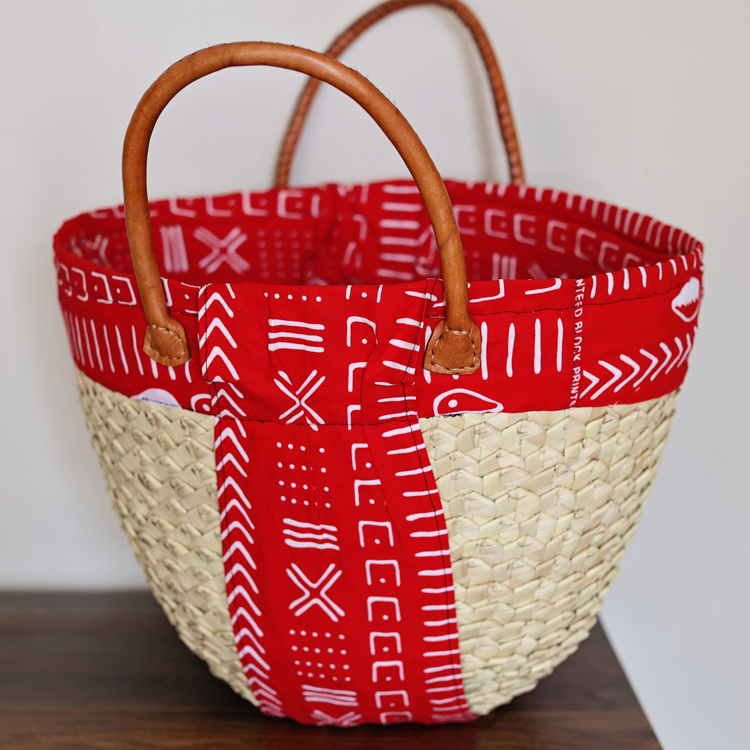 A woven straw tote bag with a red patterned fabric lining and handles, resting on a dark wooden surface against a plain light gray wall.