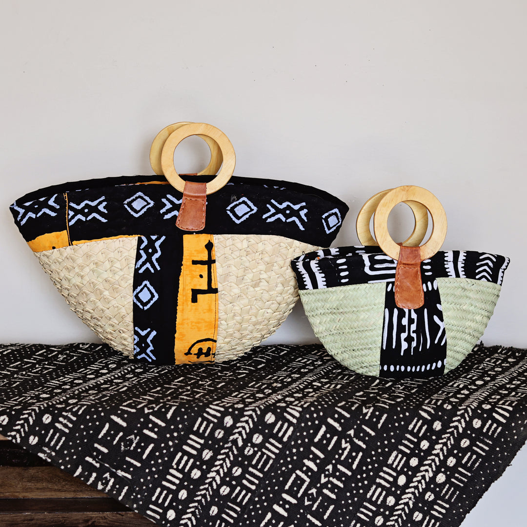 Two woven handbags with circular wooden handles are displayed on a patterned textile against a plain light wall. The larger bag is beige with black, yellow, and blue patterned panels, and the smaller bag is beige with black, yellow, and pale green patterned panels.