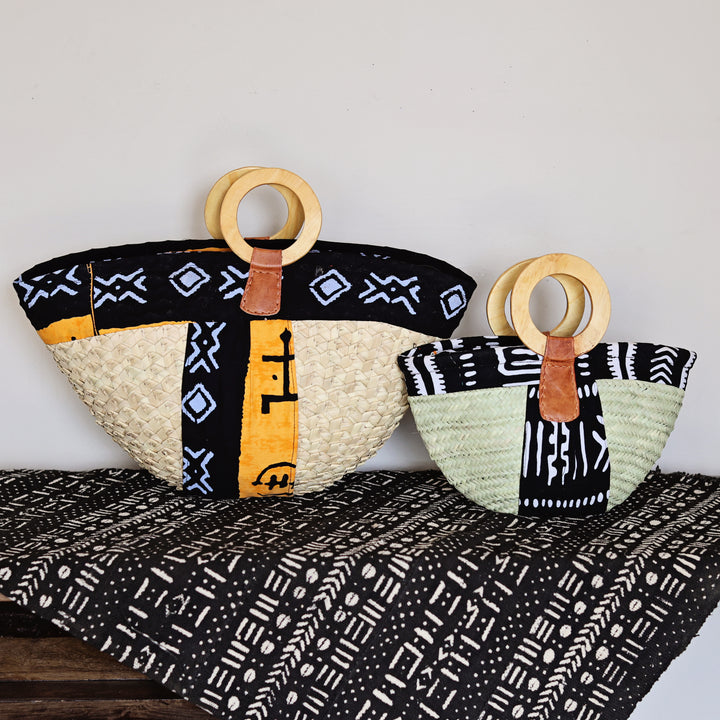 Two woven handbags with circular wooden handles are displayed on a patterned textile against a plain light wall. The larger bag is beige with black, yellow, and blue patterned panels, and the smaller bag is beige with black, yellow, and pale green patterned panels.