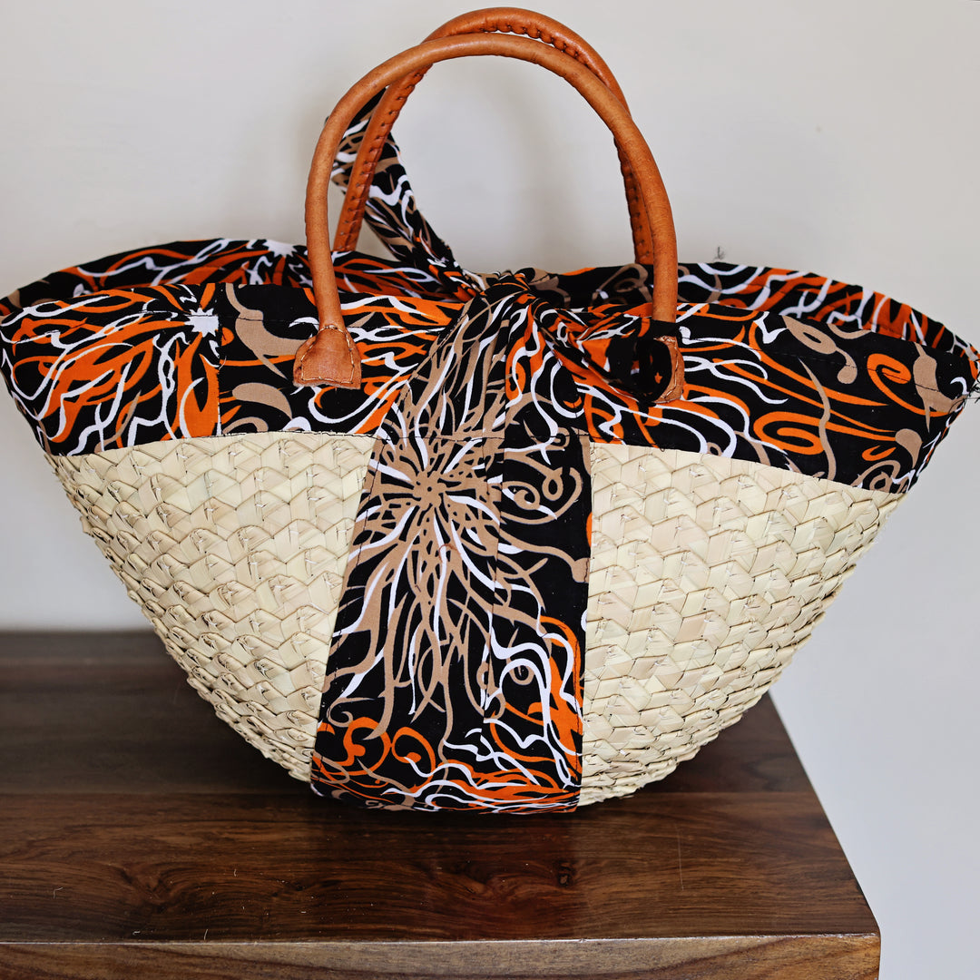 A woven tote bag with a natural straw body, brown leather handles, and a black fabric body featuring orange, beige, and white abstract swirl patterns, resting on a dark wooden surface against a plain light background.