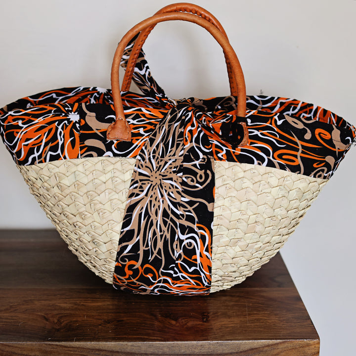 A woven tote bag with a natural straw body, brown leather handles, and a black fabric body featuring orange, beige, and white abstract swirl patterns, resting on a dark wooden surface against a plain light background.