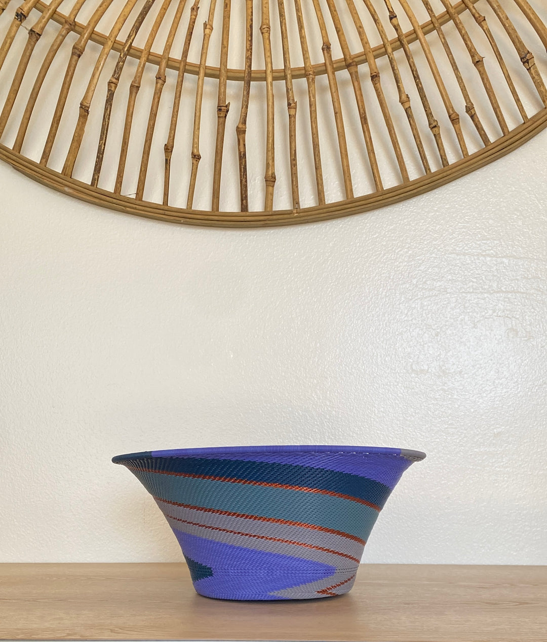 A handwoven large flared purple zulu bowl with a blue, teal, and gray pattern and thin orange-red stripes, sitting on a light wood surface against a white wall with a bamboo wall hanging above.