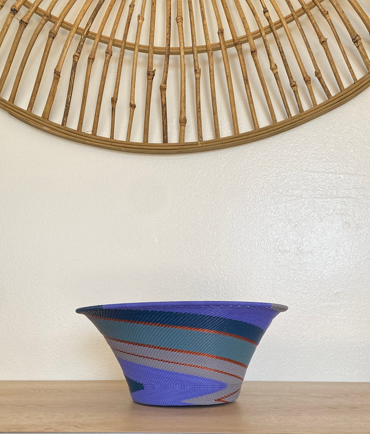 A handwoven large flared purple zulu bowl with a blue, teal, and gray pattern and thin orange-red stripes, sitting on a light wood surface against a white wall with a bamboo wall hanging above.