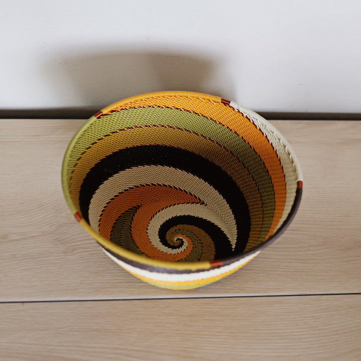 A hand-woven zulu basket bowl with a spiral pattern of yellow, orange, black, beige, and olive green, placed on a light wood surface against a white wall.