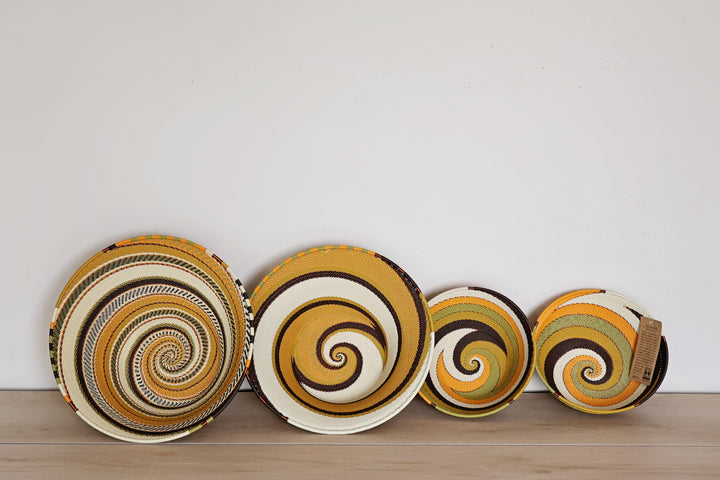 Four light earth zulu plates with colorful spiral patterns in yellow, brown, white, and green, arranged in descending size order on a light wooden surface against a plain white wall.