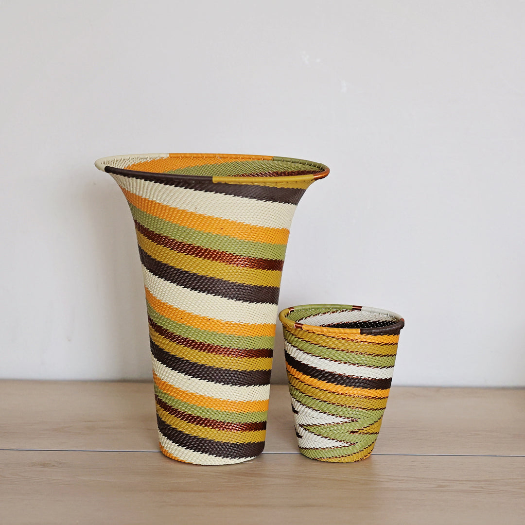 Two hand-woven vase zulu basket vases with a striped pattern in orange, yellow, green, brown, and cream, displayed on a light wood surface against a plain white wall.