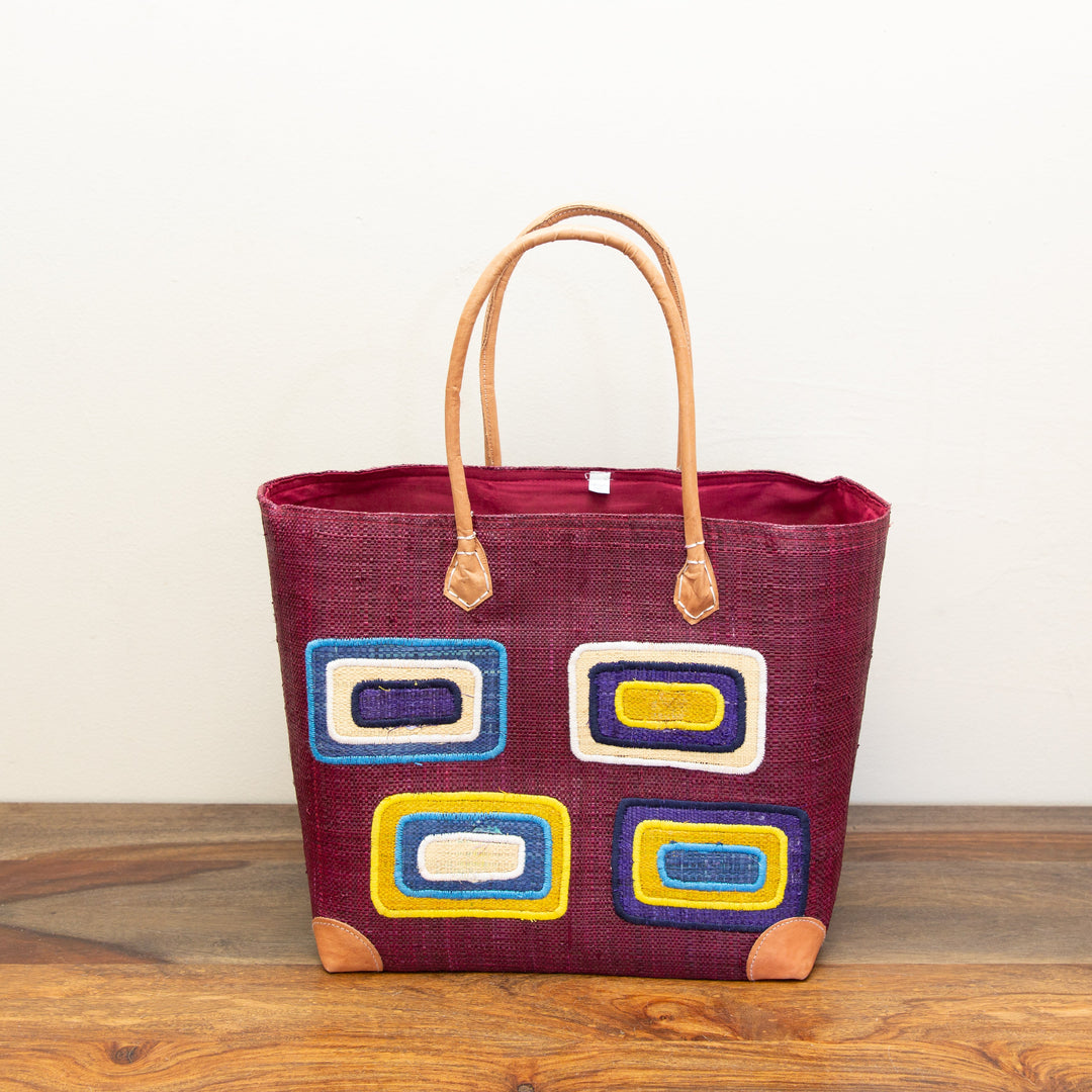 Maroon tote bag with colorful, geometric square patterns on a wooden surface.