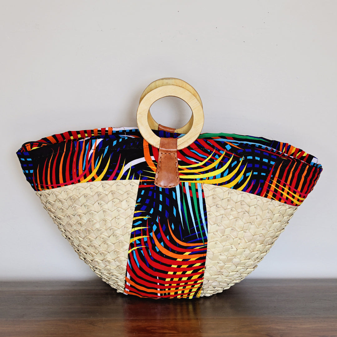 A woven straw tote bag with a colorful African wax print panel and a circular wooden handle, resting on a dark wooden surface against a plain white wall.