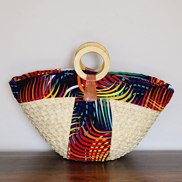 A woven straw tote bag with a colorful African wax print panel and a circular wooden handle, resting on a dark wooden surface against a plain white wall.
