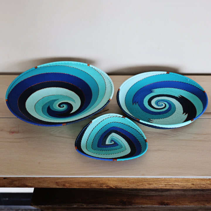 Three hand-woven decorative zulu baskets in Ocean with spiral patterns in shades of blue, teal, black, and light aqua, arranged on a light wood table against a plain wall.