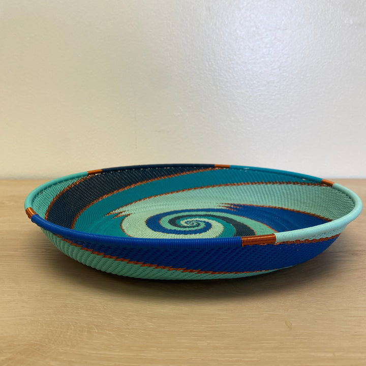 A hand-woven Ocean Zulu round basket large tray with a spiral pattern in shades of blue, teal, and light green, accented with orange and dark blue stripes, resting on a light wood surface against a plain beige wall.