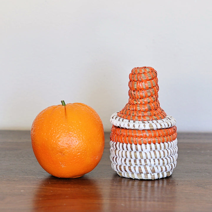 Fair-trade, mini orange and white sweetgrass basket with lid, positioned next to an orange fruit to show the true size.