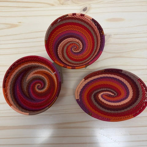 Three small woven zulu spiral-patterned bowls in warm red, orange, purple, and brown tones, arranged on a light wood surface.