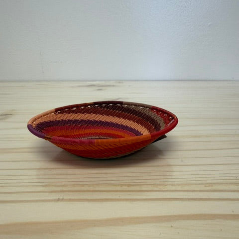 A small hand-woven zulu basket bowl with a striped pattern in red, orange, brown, and purple, resting on a light wood tabletop against a white wall.